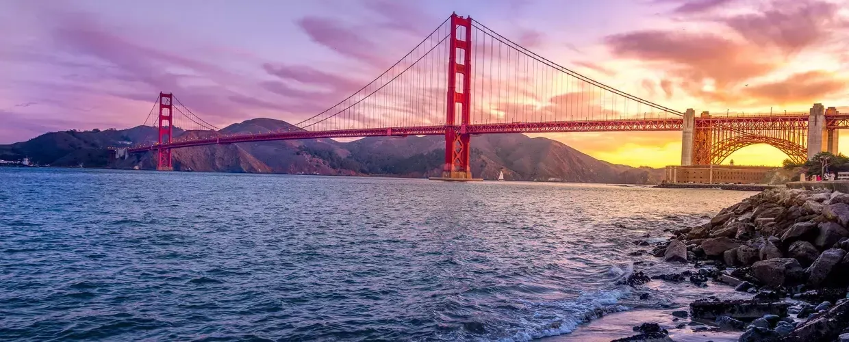 的 金门大桥 at sunset with a multicolored sky and the San Francisco Bay in the foreground.